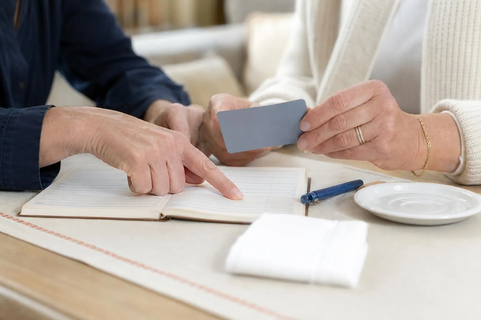 Deux mains de femmes au-dessus d’une table basse où sont posés un carnet de rendez vous ouvert, une carte bancaire neutre et un stylo